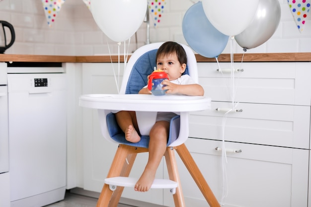 Photo little baby boy sitting in high chair at home on white kitchen and drinking water from sippy cup on background with balloons.