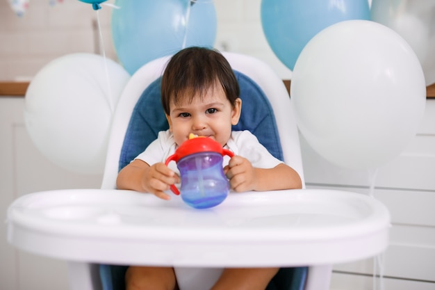 Little baby boy sitting in high chair at home on white kitchen and drinking water from sippy cup on background with balloons.