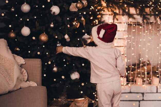 Little baby decorate Christmas tree wearing pajamas and red santa claus hat