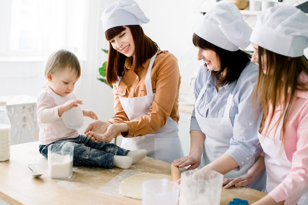 Little baby girl is sitting on the wooden table at kitchen and having fun with sugar. Grandmother and her daughters are baking cookies. Happy women in white aprons baking together. Mothers Day