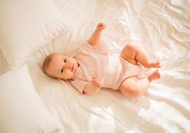 A little baby girl in a pink bodysuit is lying on the bed