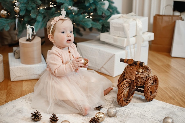 Little baby girl sitting on the floor near christmas tree at home. Girl is playing with balls for Christmas tree decoration. Girl wearing peach dress.