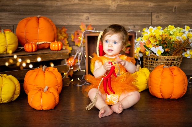 Little baby girl sitting with pumpkins in a bright orange dress on a brown background, halloween