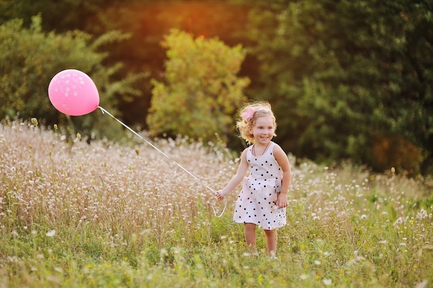 Little baby girl in white dress with pink air balloon on a background of greenery, grass at sunset.