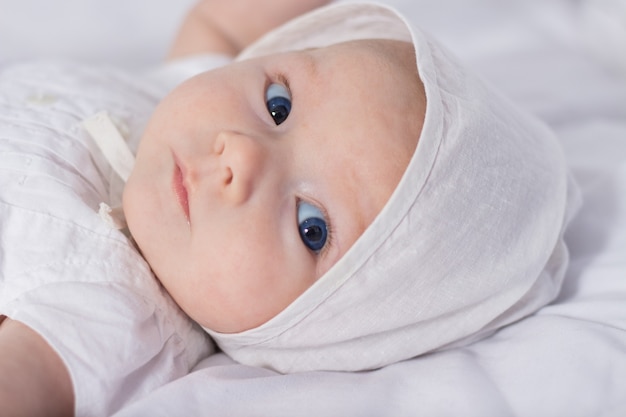 Little baby girl with blue eyes in white dress and hat on white blanket.
