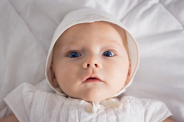 Little baby girl with blue eyes in white dress and hat on white blanket.