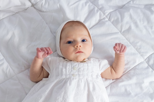 Little baby girl with blue eyes in white dress on white blanket.