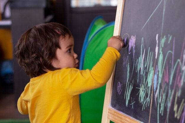 A little  baby girl in a yellow sweatshirt draws on a blackboard with colored crayons