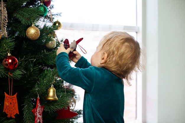 小さな赤ちゃんがクリスマスツリーにおもちゃを掛けます。家で新年とクリスマス。冬休み。クリスマスツリーを飾ります。