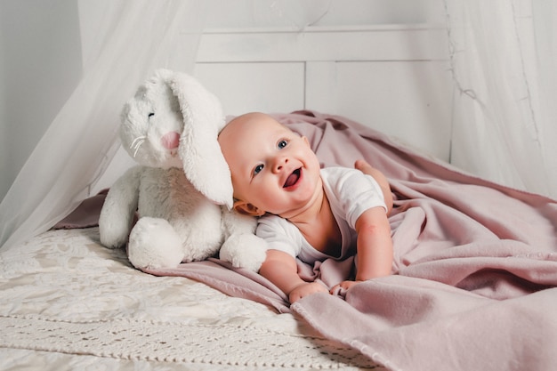 A little baby lays on a bed with a toy rabbit and smiles