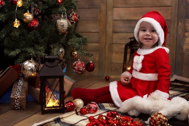 Little baby sitting on the floor in the room in front of a decorated Christmas tree