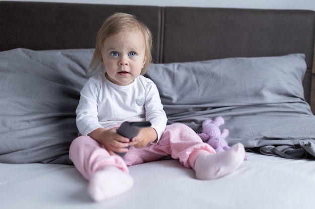 Little baby toddler with blue eyes sits on the bed in the\
bedroom with a tv remote control