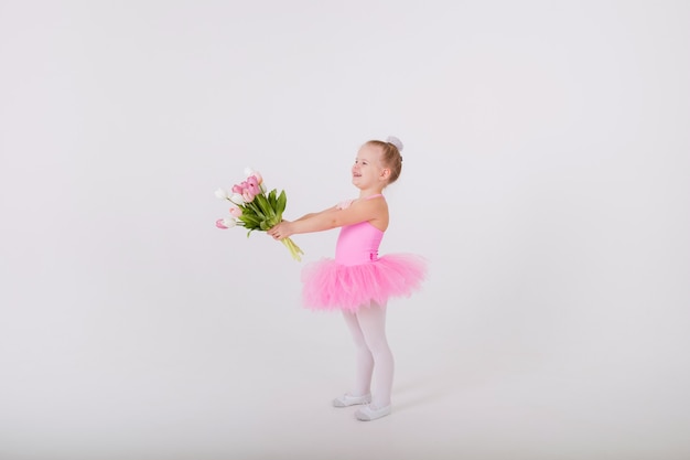 Little ballerina girl in a tutu skirt presents a bouquet of colorful tulips on a white wall