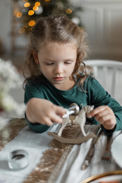 녹색 패셔너블한 옷을 입은 작고 아름다운 여자 아이는 축제 테이블에 앉아 크리스마스 이브에 목마 장난감을 가지고 논다