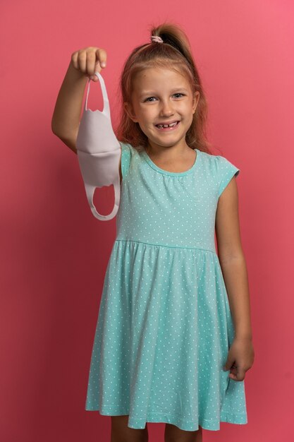 Little beautiful child girl in blue dress take off face mask\
and smile on pink background