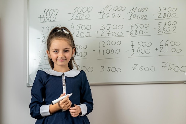 Little beautiful girl answers in front of the school blackboard smiling