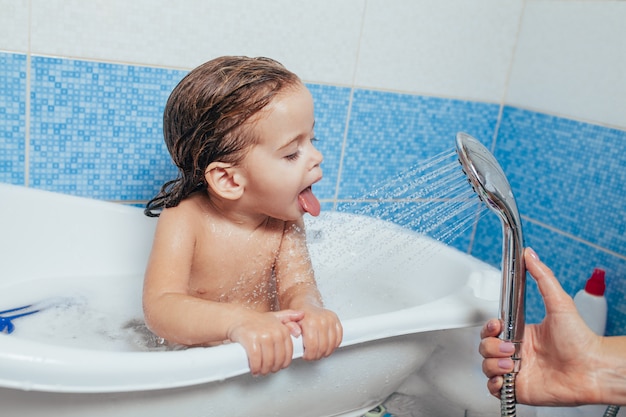 Little beautiful girl bathes in a bathroom, plays the fool and shows tongue.