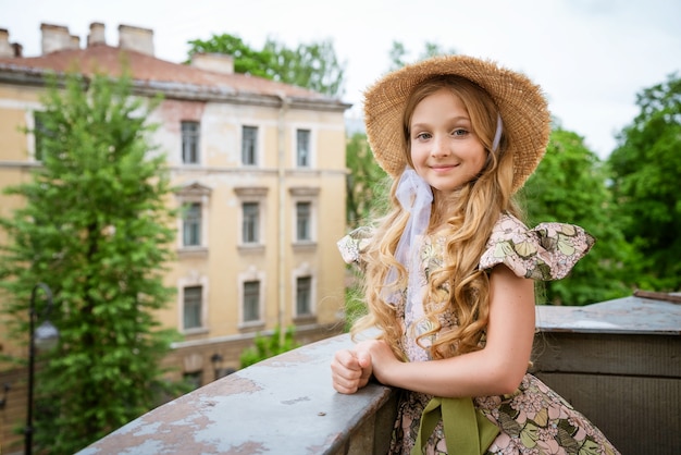 Маленькая красивая девушка в платье и шляпе позирует на балконе на фоне городской пейзаж