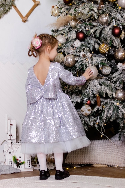 A little beautiful girl in an elegant dress with her back turned stands near the Christmas tree