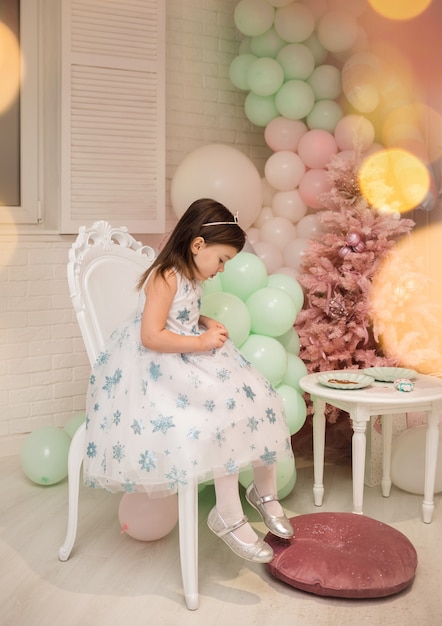 A little beautiful girl in a fluffy white dress is sitting on a chair in the New Year decorations