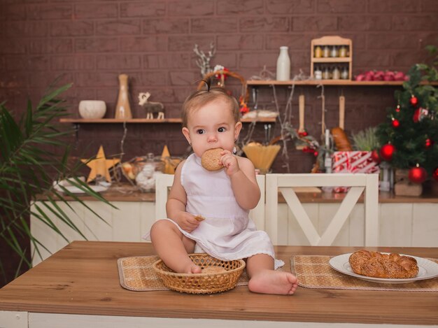 Photo a little beautiful girl is sitting on the kitchen table in a cooks costume and eating cookies