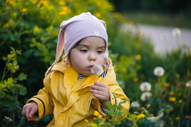 Little beautiful girl in a pink cap and a yellow jacket sits in a field of yellow dandelions and flowers