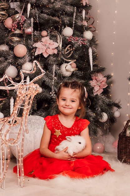 Little beautiful girl smiles and holds a white fluffy rabbit in her arms near the Christmas tree