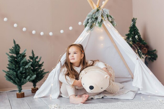 Foto una bambina bellissima in pigiama bianco gioca con un orsacchiotto vicino alla stanza dei bambini del teepee con ca