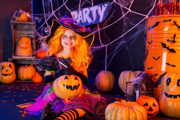 Little beautiful girl in a witch costume celebrates Happy Halloween party in interior with pumpkins.