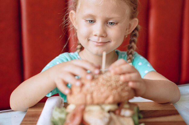 Little beautiful girl with blond hair is looking with an appetite for a juicy huge burger in a cafe place. Portrait.