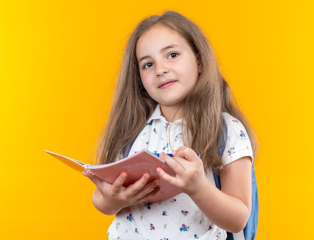 Little beautiful girl with long hair with backpack holding notebook looking at front smiling confident standing over orange wall