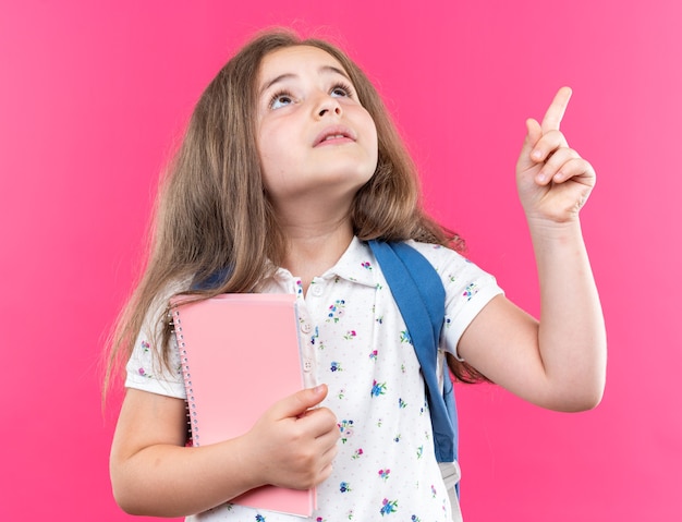 Little beautiful girl with long hair with backpack holding notebook looking up intrigued pointing with index finger up standing over pink wall