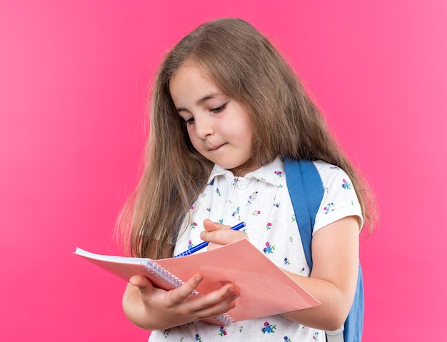 Piccola bella ragazza con i capelli lunghi con lo zaino che tiene il taccuino che scrive qualcosa con la penna che sembra sicura in piedi sul rosa