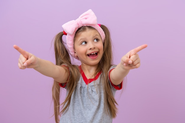 A little beautiful girl with a pink bow on her head smiles and points her fingers to the side.