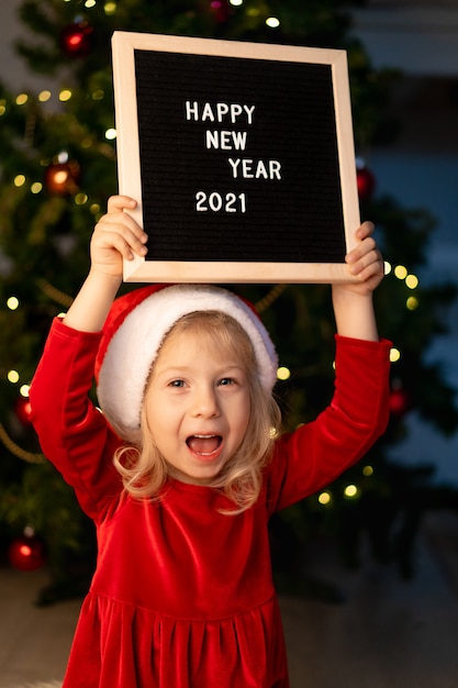 Little beautiful girl with Santa Claus hat and red Christmas dress is lying under the Christmas tree and laughing, next to a black Board and white letters text happy new year 2021. High quality photo