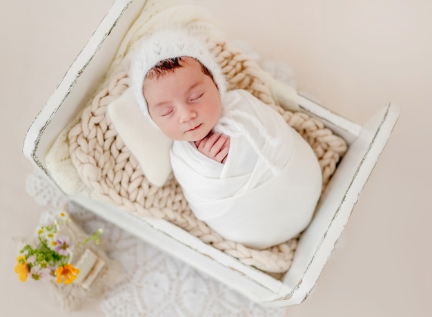 Piccola bellissima neonata fasciata in tessuto e con indosso un cappello lavorato a maglia che dorme in un piccolo letto progettato durante il servizio fotografico in studio. simpatico bambino neonato che fa un pisolino a riposo