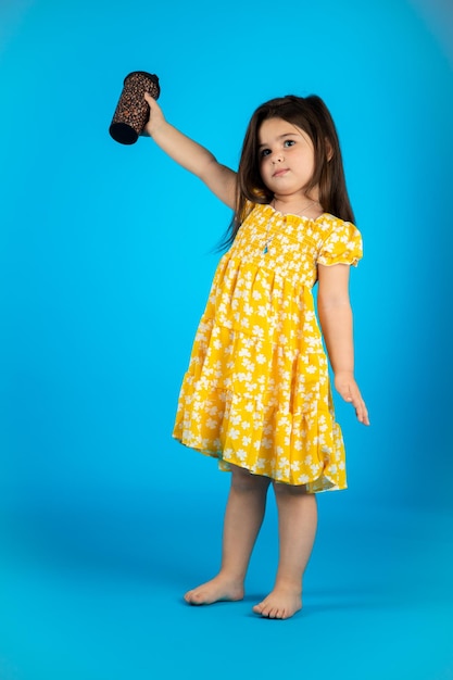 Little beautiful smiling girl with a funny face in a striped yellow dress posing on a blue background in studio