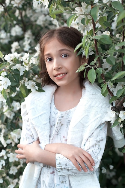 A little beautiful teenage girl in a white dress and sweater enjoys the spring flowering of apple trees smiles and looks at the camera Spring