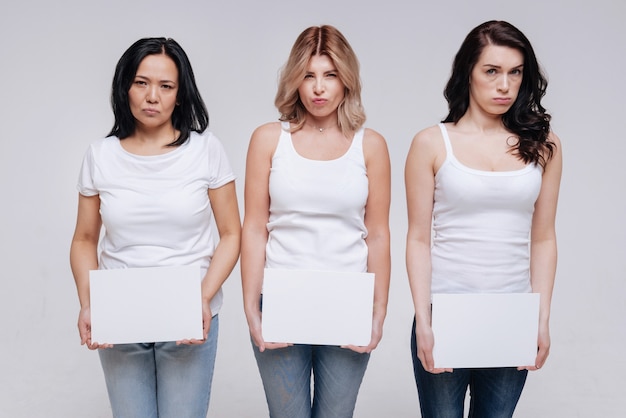 Little bit upset. Artistic pretty stunning ladies acting on set while holding white pieces of paper and standing close to each other