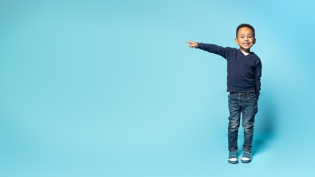 Little black boy pointing aside at free space on blue studio background demonstrating free place for