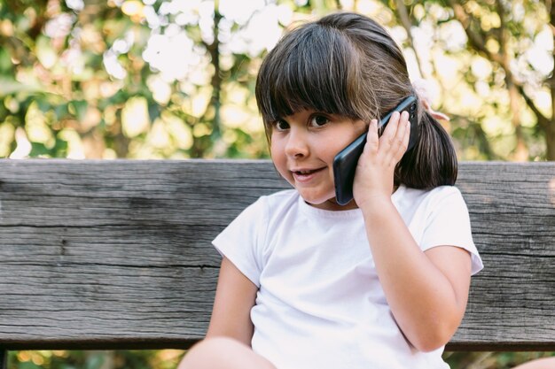 Little black-haired girl, wearing a white t-shirt, sitting on a park bench talking on the mobile phone, smiling.