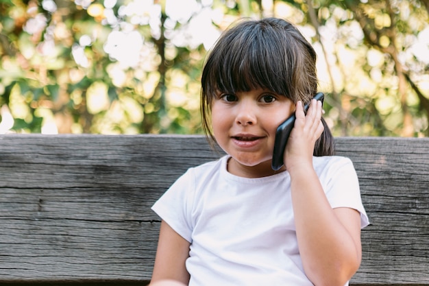 Little black-haired girl, wearing a white t-shirt, sitting on a park bench talking on the mobile phone, smiling.