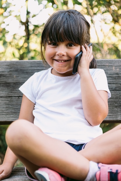 Маленькая черноволосая девочка в белой футболке сидит на скамейке в парке, разговаривает по мобильному телефону и улыбается.