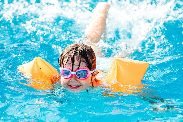 Piccola ragazza dai capelli neri sorridente e nuotare in piscina indossando maniche e occhiali da nuoto