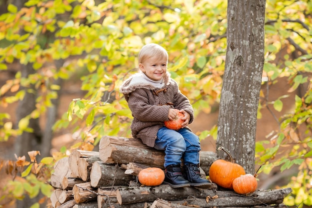 A little blond boy in autumn clothes is smiling sitting in the forest on logs with pumpkins