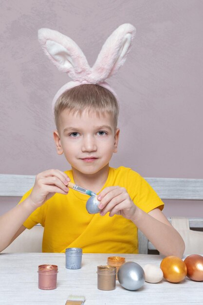 A little blond boy in bunny ears paints eggs for the Easter holiday in the home kitchen vertical frame The child has fun and celebrates the holiday DIY Easter eggs concept