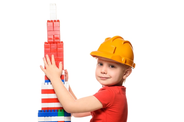 Маленький белокурый мальчик в строительном шлеме и красной рубашке стоит возле башни, построенной из деталей конструктора. Портрет. Изолировать на белом фоне.