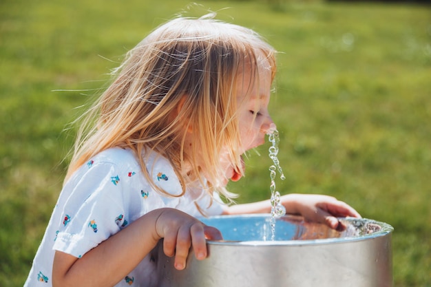 A little blond boy drinks from a drinking fountain in a city park on a hot summer day outdoors