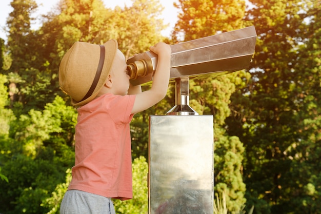 Photo little blond boy in hat looking in a large pair of binoculars.