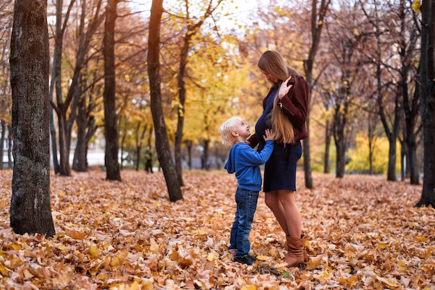 Little blond boy hugs his pregnant mom's belly. Autumn park on the background